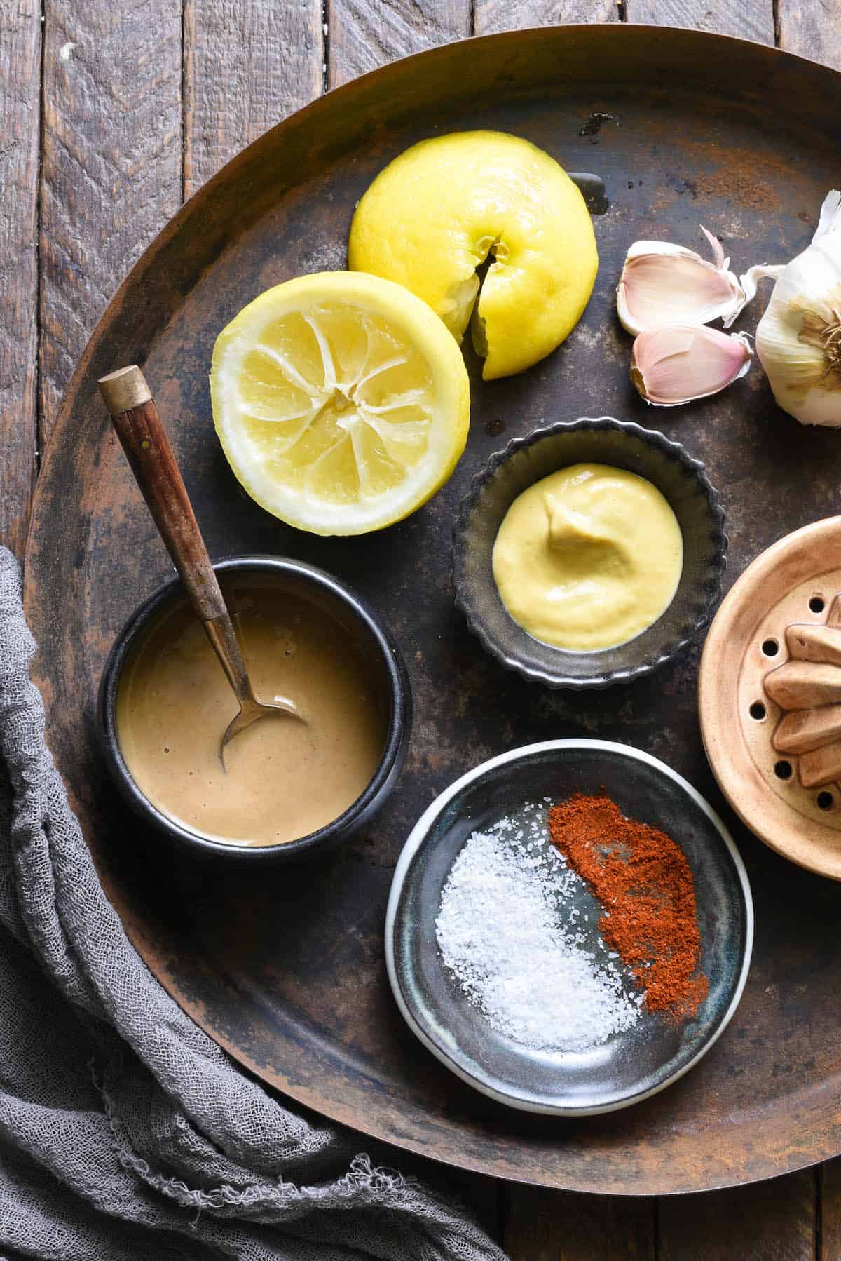 Overhead shot of ingredients for lemon tahini dressing. Tahini, salt and paprika, dijon mustard, lemon halves and garlic cloves.