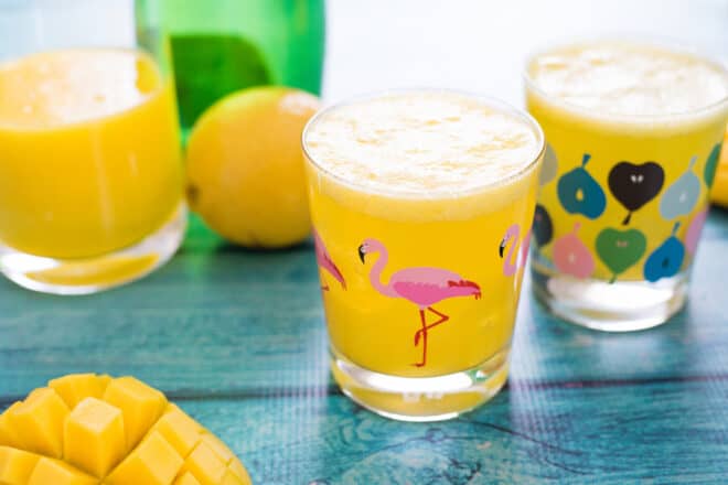 Two glasses of sparkling mango drink on teal table, with fruit and bottles of juice and water in background and foreground.