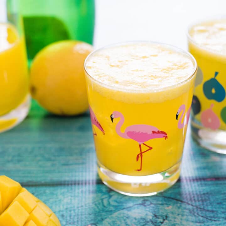 Two glasses of sparkling mango drink on teal table, with fruit and bottles of juice and water in background and foreground.