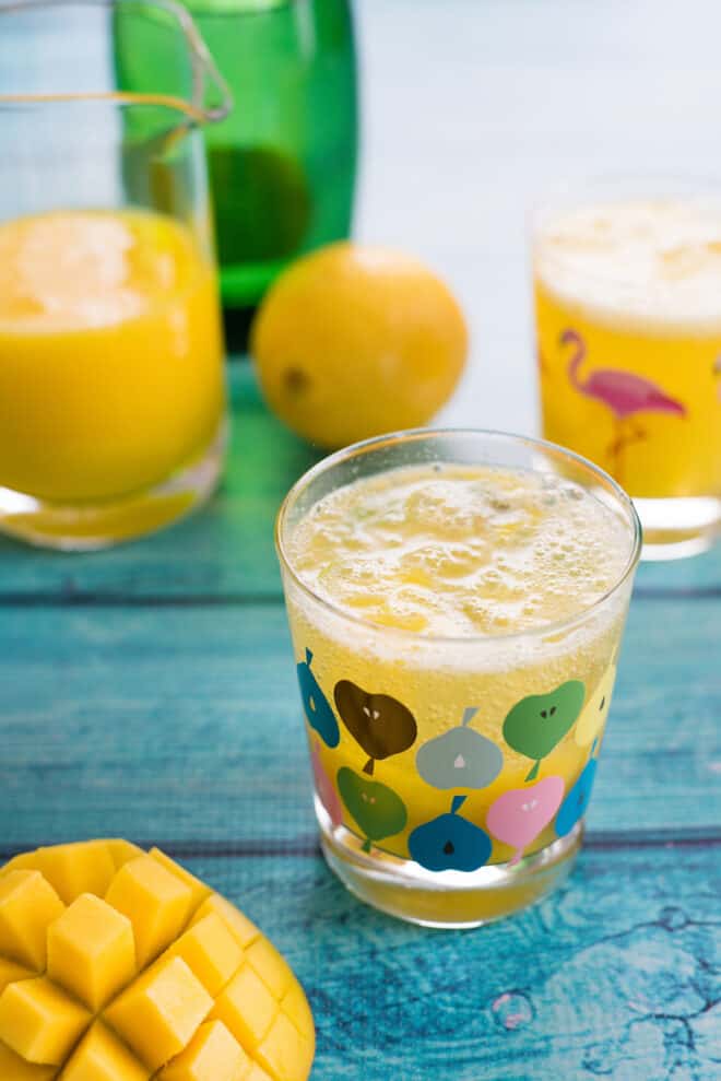 Two glasses of mango lemonade on teal table, with fruit and bottles of juice and water in background and foreground.