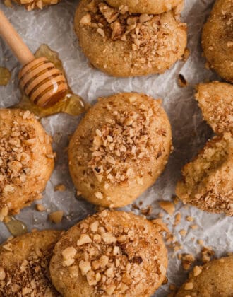 Greek cookies sprinkled with walnuts on white parchment paper with a honey dipper.