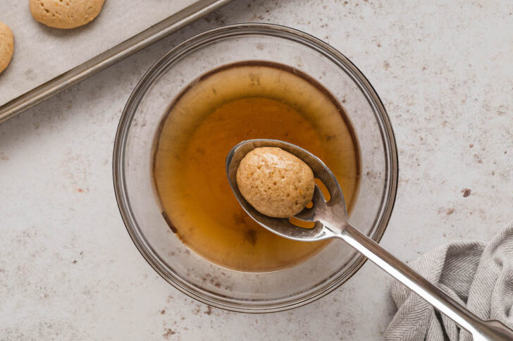 A slotted spoon dipping a light brow oval-shaped cookie into a syrup mixture.