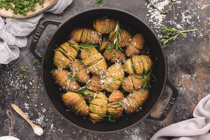 Mini Hasselback potatoes in a cast iron skillet, sprinkled with rosemary sprigs and shredded Parmesan cheese.