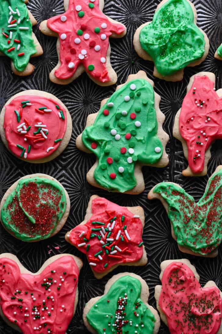 Frosted sour cream cookies with sprinkles in a variety of shapes on a textured baking pan.