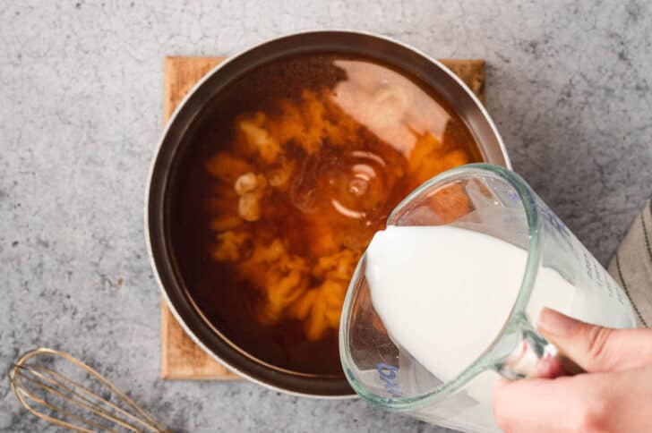 A stainless steel saucepot filled with brown broth, with a hand pouring in a white liquid from a measuring cup.