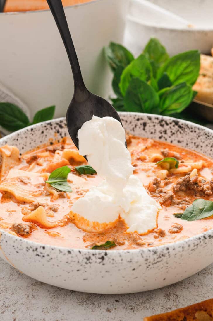 A bowl of pasta and ground beef soup being dolloped with a spoonful of ricotta cheese.