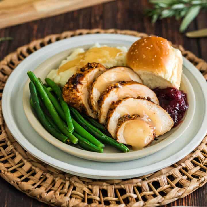 A turkey tenderloin recipe, sliced, on a plate with green beans, mashed potatoes and gravy, a roll and cranberry sauce.