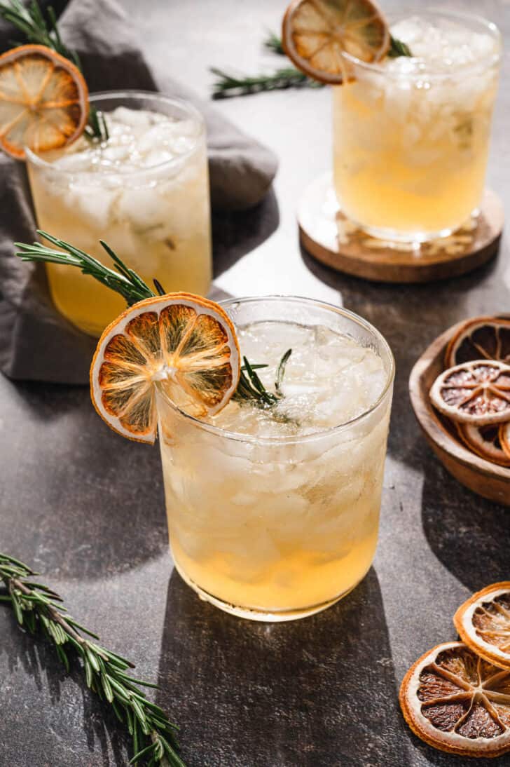 A light orange hued drink in an old fashioned glass, garnished an herb sprig and a dried citrus slice.