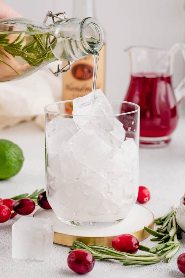 A double old fashioned glass filled with ice, with rosemary simple syrup being poured over the ice.