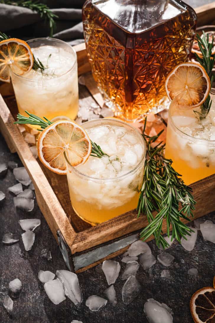 Rosemary drinks garnished with rosemary sprigs and dried citrus on a wooden tray with ice cubes all around the scene.