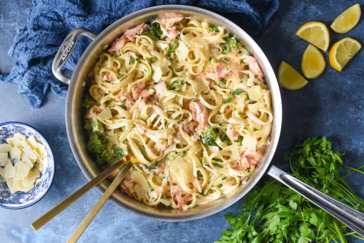 A large stainless steel skillet filled with salmon fettuccine alfredo, with Parmesan cheese, lemon wedges and parsley garnishing the scene.