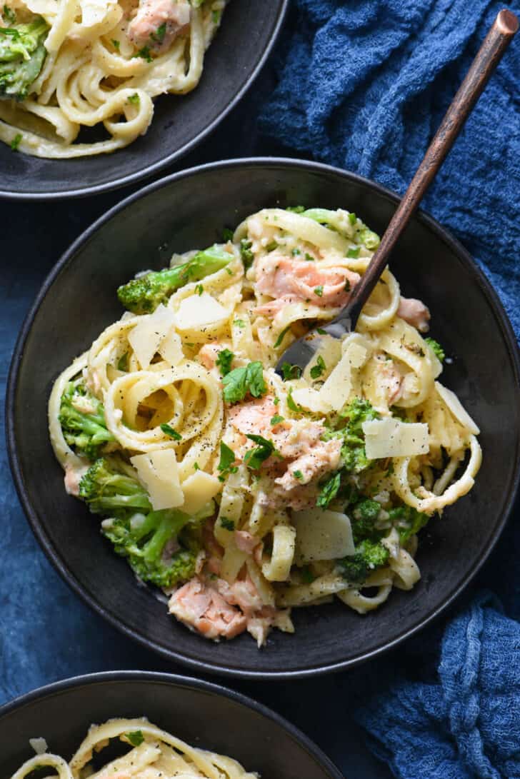 A black bowl filled with salmon fettuccine and broccoli, with a fork digging in.