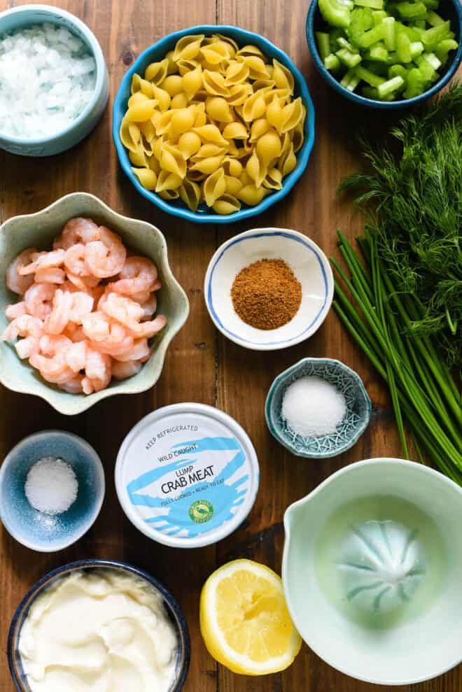 Wooden tabletop with ingredients for seafood pasta salad in small bowls, including pasta, spices, herbs, vegetables and mayonnaise.