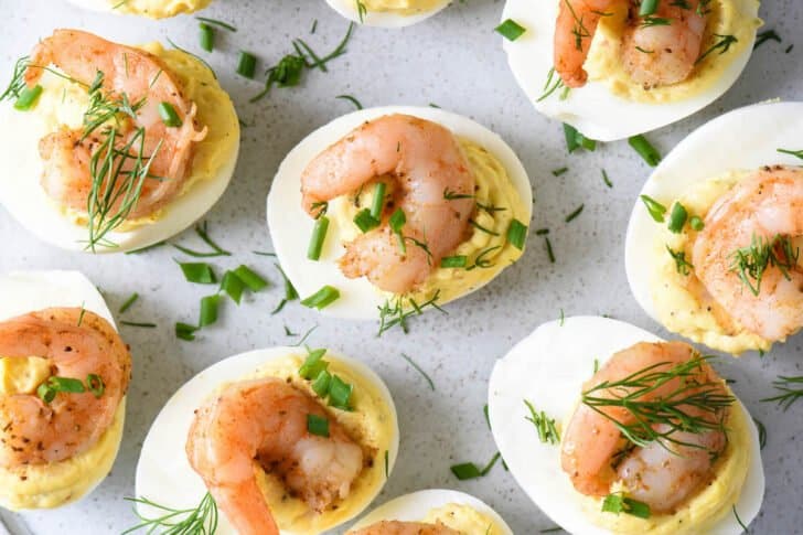 Seafood deviled eggs topped with shrimp and garnished with chives and dill on a light gray plate.