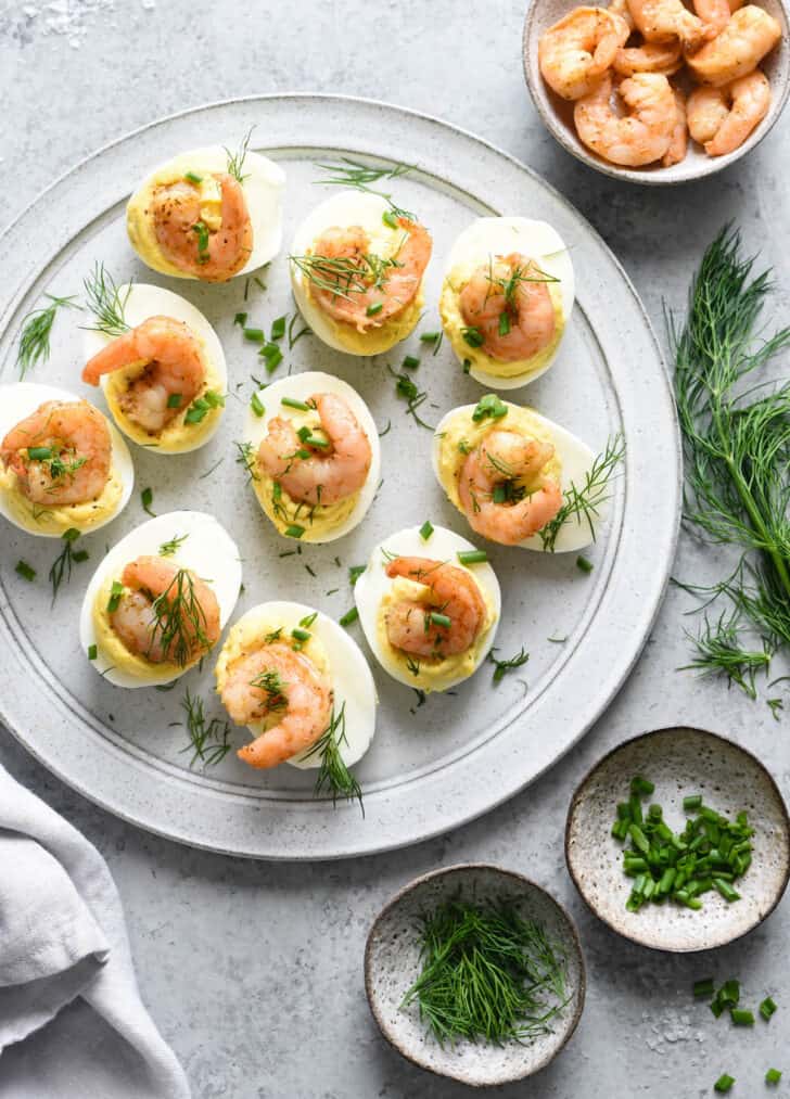 Deviled eggs with shrimp, garnished with fresh herbs, on a light gray plate, with bowls of fresh herbs and cooked shrimp nearby.