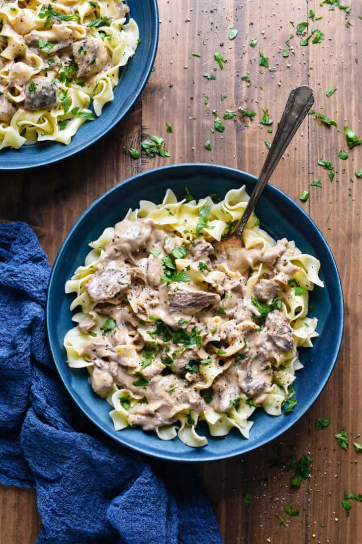 Two blue bowls filled with crock pot beef stroganoff on a wooden background with a blue napkin.