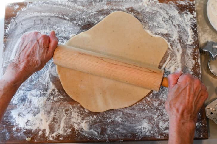 A large flour wooden cutting board with a woman's hands rolling out cookie dough on top of it with a rolling pin.