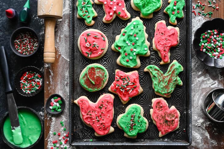 Frosted sour cream sugar cookies with sprinkles in a variety of shapes on a textured baking pan.