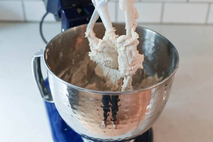 A white frosting mixture in a Kitchenaid stand mixer bowl being beat with the paddle attachment.