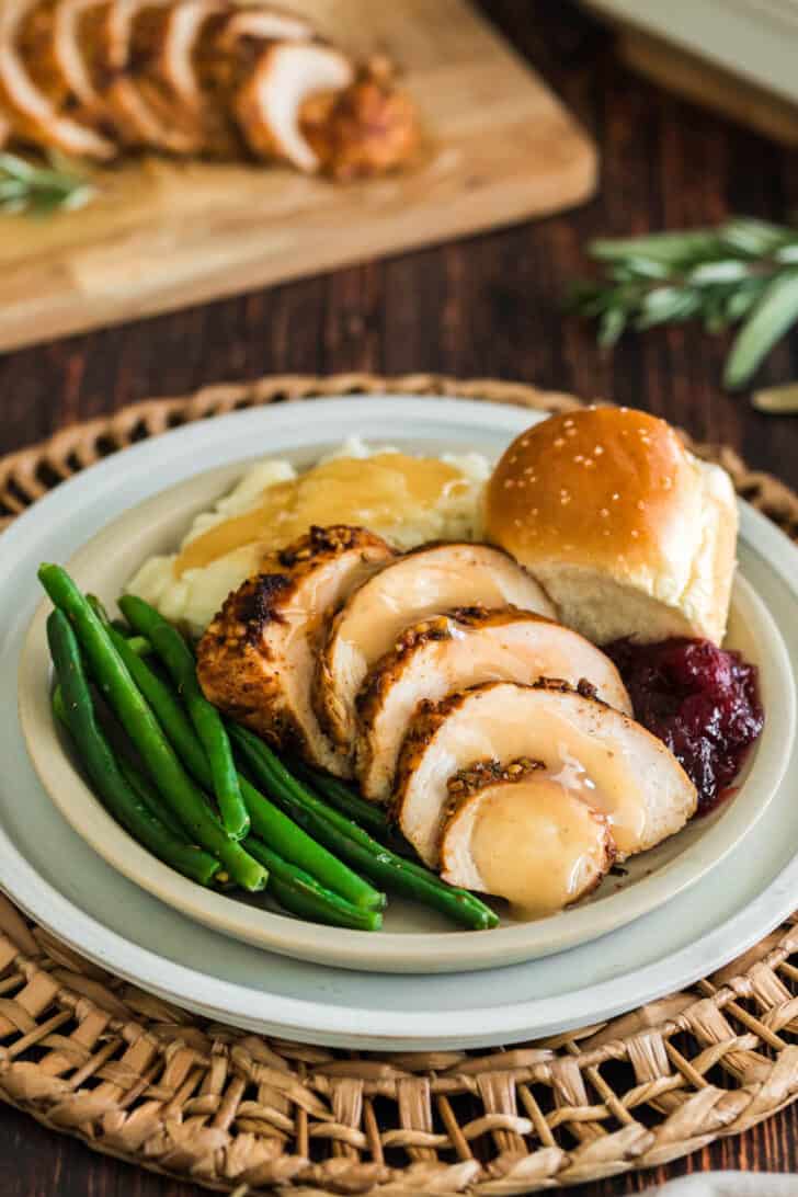 A turkey tenderloin recipe, sliced, on a plate with green beans, mashed potatoes and gravy, a roll and cranberry sauce.